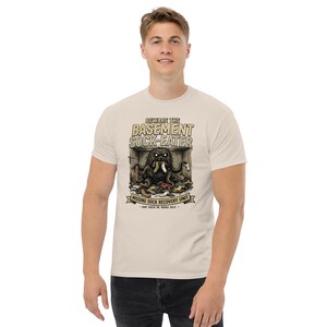 May include: A cream-colored t-shirt featuring a graphic of a monster labeled "Basement Sock-Eater." The illustration shows a creature surrounded by socks, with text reading "Missing Sock Recovery Unit" and "One sock in, none out."