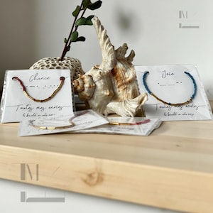 Peut inclure: Trois bracelets avec des fermoirs dorés et des perles colorées sur des cartes blanches avec les mots "Chance", "Joie" et "Toutes mes envies" écrits dessus. Les bracelets sont exposés sur une surface en bois avec une coquille.