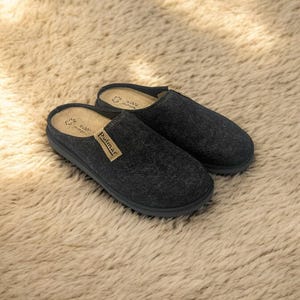 Puede incluir: Un par de pantuflas de lana gris oscuro con suela de goma negra. Las pantuflas tienen un diseño sencillo y están etiquetadas con "Pullmar" en la parte superior.