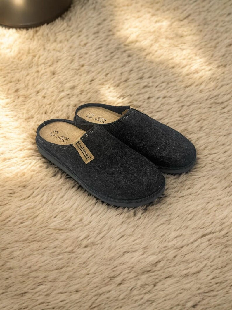 Puede incluir: Un par de pantuflas de lana gris oscuro con suela de goma negra. Las pantuflas tienen un dise&ntilde;o sencillo y est&aacute;n etiquetadas con "Pullmar" en la parte superior.