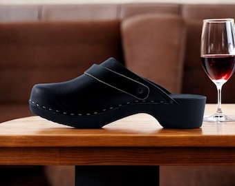 Op de afbeelding: Een zwarte leren klomp met een houten zool en een band over de bovenkant. De klomp staat op een houten tafel naast een glas rode wijn.