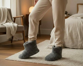 May include: A pair of charcoal gray ankle-high slippers made of a textured, fleece-like material. The slippers feature a rounded toe, a thick sole, and a simple pull-on design that covers the foot and ankle, worn with beige sweatpants.