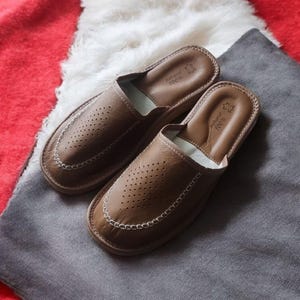 Op de afbeelding: Een paar bruine leren slippers met een geperforeerd ontwerp en een witte voering. De slippers hebben een gestikte rand en liggen op een grijs oppervlak.