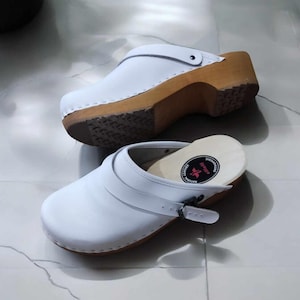 May include: A pair of white leather clogs with a wooden sole and a buckle strap. The clogs have a red and black logo on the insole.