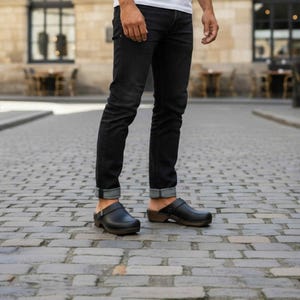 Könnte beinhalten: Ein Paar schwarze Leder-Clogs mit Holzsohle und silbernem Schnallenriemen, getragen zu einer schmal geschnittenen, dunkelblauen Jeans mit umgeschlagenem Saum. Das Schuhwerk hat eine abgerundete Kappe und eine robuste Holzsohle.