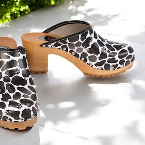 Peut inclure: Une paire de sabots à talon en bois avec un imprimé animal noir et blanc. Les sabots sont ornés de clous et sont en cuir.