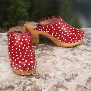 Könnte beinhalten: Ein Paar rote Clogs mit weißen Punkten und Holzabsätzen. Die Clogs haben ein weißes Leder-Obermaterial und eine Holzsohle.