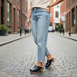 May include: A pair of light blue denim jeans with a high-waisted fit and straight legs, paired with black leather clogs featuring wooden soles and silver-toned metal studs along the sides, worn with a plain beige short-sleeved t-shirt.
