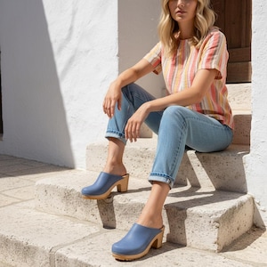 Pode incluir: Um par de tamancos de couro azul com saltos e solas de madeira. Os tamancos apresentam uma biqueira arredondada, uma superfície superior lisa e um salto bloco resistente. Estes sapatos sem atacadores têm uma base de madeira natural.
