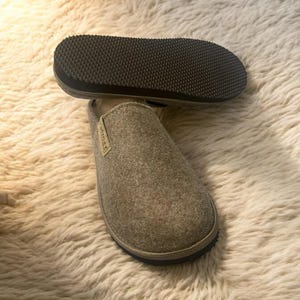 Puede incluir: Un par de zapatillas de fieltro gris con suelas negras. Las zapatillas est&aacute;n sobre una alfombra blanca y esponjosa.