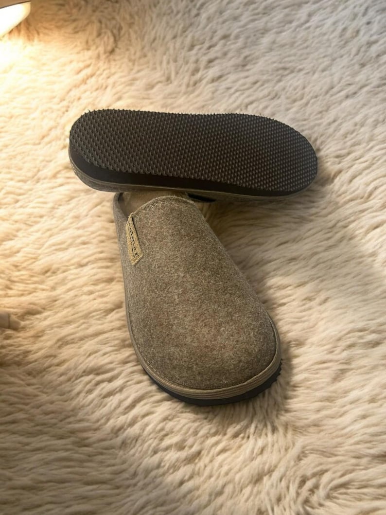 Puede incluir: Un par de zapatillas de fieltro gris con suelas negras. Las zapatillas est&aacute;n sobre una alfombra blanca y esponjosa.