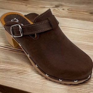 Pode incluir: Sapatilha de couro marrom com uma fivela e um salto de madeira. A sapatilha tem uma sola cravejada.