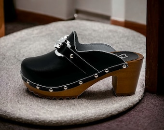 Op de afbeelding: Zwarte leren klomp met een houten hak en een zilveren gesp. De klomp heeft een wit stikseldetail en zilveren studs rond de rand.