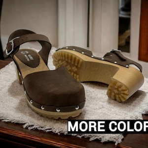 Può includere: Due paia di zoccoli in pelle marrone con chiusura a fibbia. Gli zoccoli hanno un tacco in legno e una suola bianca. Gli zoccoli sono su un asciugamano bianco.