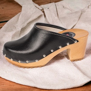 Puede incluir: Zueco de cuero negro con tacón de madera y detalles de tachuelas. El zueco está sobre una superficie de tela blanca.