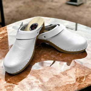 Może przedstawiać: Para białych drewnianych klapek z paskiem z klamrą. Klapki leżą na marmurowej powierzchni.