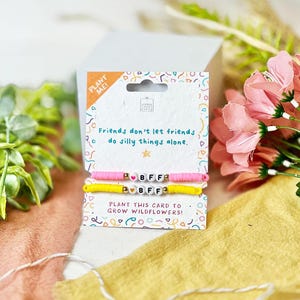 Könnte beinhalten: Zwei Freundschaftsarmbänder mit den Buchstaben "BFF" auf einer Karte mit dem Text "Freunde lassen Freunde keine dummen Dinge allein tun." Ein Armband ist rosa, das andere gelb. Auf der Karte steht "Pflanze diese Karte, um Wildblumen anzubauen!"