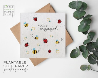 May include: A square, off-white engagement card with the words "you're engaged!" surrounded by illustrations of bees, ladybugs, and engagement rings. The card is made of plantable seed paper and rests on a brown envelope.