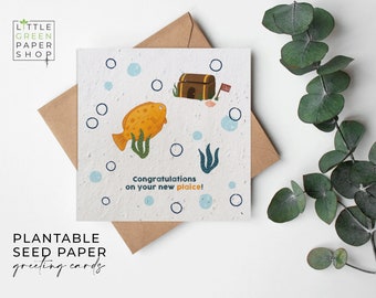 Può includere: Un biglietto di auguri in carta da seme piantabile con un'illustrazione di un pesce dei cartoni animati che nuota vicino a uno scrigno del tesoro con una bandiera "Venduto". La carta ha uno sfondo blu e bianco con il testo "Congratulazioni per il tuo nuovo posto!"