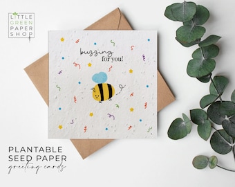 Può includere: Un biglietto d'auguri in carta da seme piantabile bianca con un'ape dei cartoni animati e il testo "buzzing for you!" circondato da coriandoli colorati.