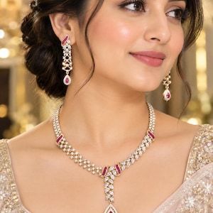 May include: A gold-toned necklace and matching drop earrings set featuring clear white stones and vibrant pink teardrop-shaped stones. The necklace has a central pendant with a pink stone, while the earrings feature a similar drop design.
