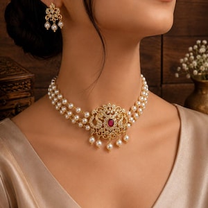 May include: A gold-toned choker necklace featuring three strands of white pearls and a central ornate pendant with a deep red oval stone, clear crystals, and dangling pearls. Matching drop earrings with a similar gold, crystal, and pearl design.