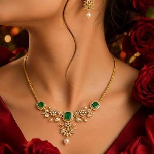 May include: A gold necklace and matching earring set featuring rectangular green stones, small white stones, and dangling white pearls. The necklace has a central green stone with floral-inspired accents, while the earring features a single green stone.