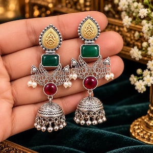May include: A pair of silver-toned dangle earrings featuring a gold-toned teardrop top, a rectangular green stone, a central red stone, and a bell-shaped jhumka base with small dangling silver beads and white pearl-like accents.