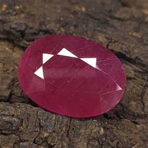 Puede incluir: Una piedra preciosa de color magenta intenso, tallada en forma ovalada con una superficie facetada. La piedra presenta un acabado pulido y liso con inclusiones internas visibles y un patrón de reflejos triangulares blanco brillante.