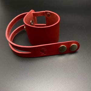 May include: Red leather cuff bracelet with two straps and silver-toned snaps. The bracelet has a small logo on the strap.