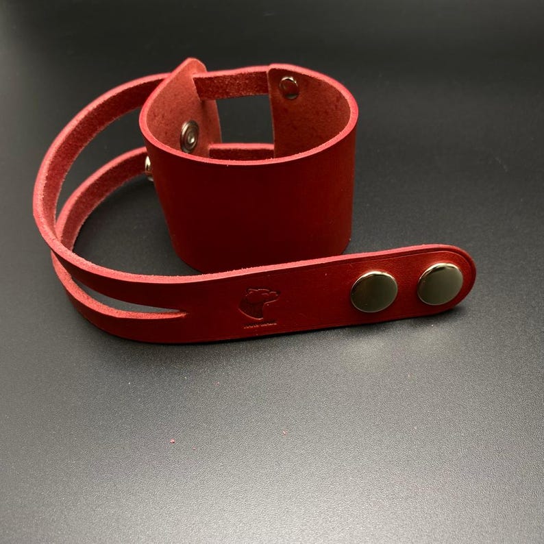 May include: Red leather cuff bracelet with two straps and silver-toned snaps. The bracelet has a small logo on the strap.