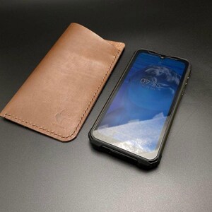 May include: A brown leather phone case with a stitched edge and a small bear logo. A black rugged phone case with a screen protector is next to it. The phone screen shows the time 09:55.