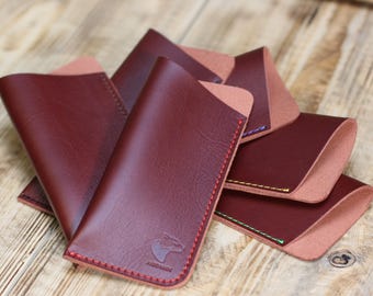 May include: Several burgundy leather eyeglass cases with contrasting stitching in red, green, and yellow. Each case has a light brown interior and a small embossed bear logo. The cases are arranged on a wooden surface.
