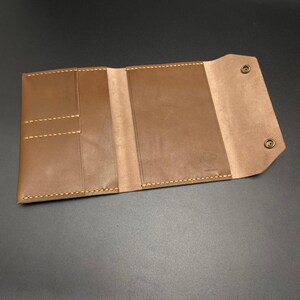 May include: A brown leather wallet with multiple pockets and a snap closure. The wallet has a stitched edge and a small logo on the front.