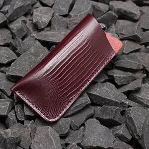 May include: A burgundy leather glasses case with a textured, embossed pattern. The case has a stitched edge and is lying on a bed of grey rocks.