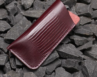 May include: A burgundy leather glasses case with a textured, embossed pattern. The case has a stitched edge and is lying on a bed of gray rocks.
