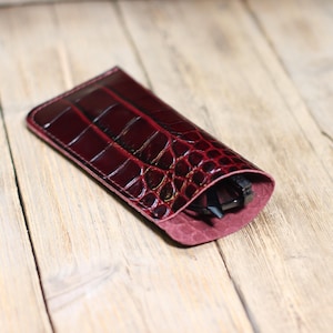 Könnte beinhalten: Ein weinrotes Lederetui für Brillen mit Krokodilhautstruktur. Das Etui ist geöffnet und zeigt eine Brille im Inneren. Das Etui liegt auf einer hellbraunen Holzoberfläche.