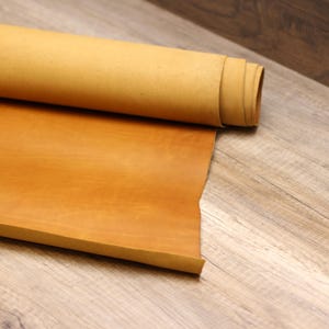 Puede incluir: Un rollo de cuero marrón claro sobre una superficie de madera. El cuero es liso y tiene un ligero brillo.