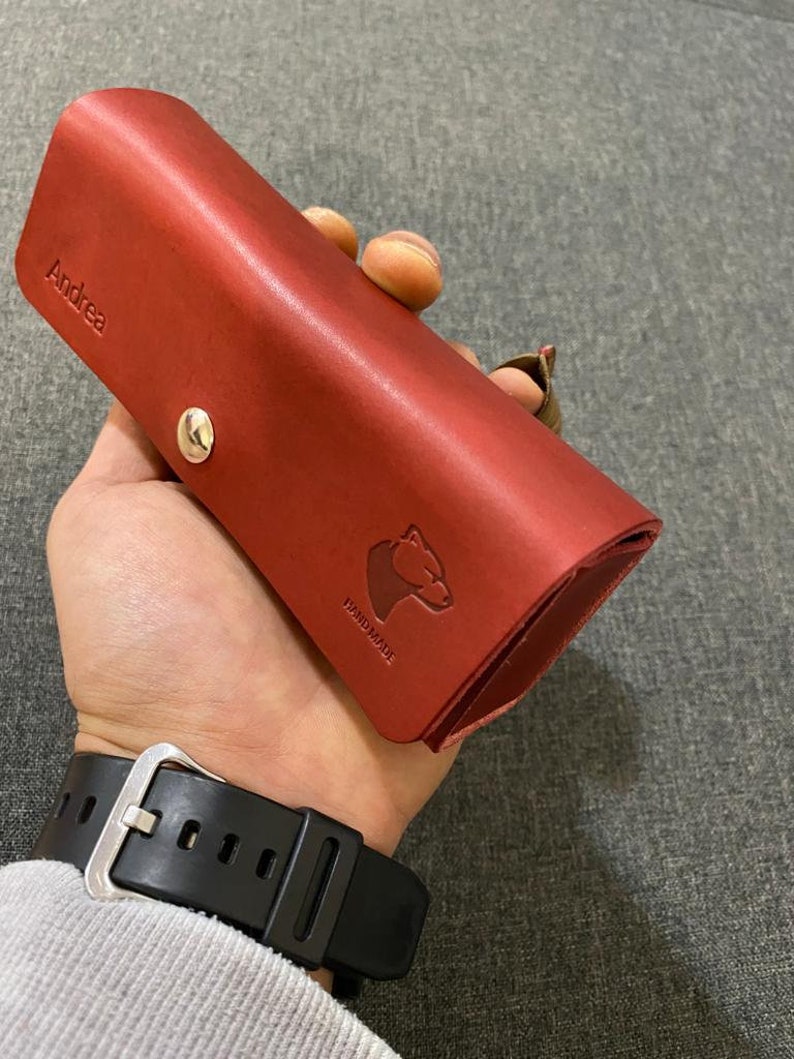 May include: A red leather case with the name "Andrea" embossed on the front. The case has a snap closure and a small bear logo with the word "Handmade" underneath.