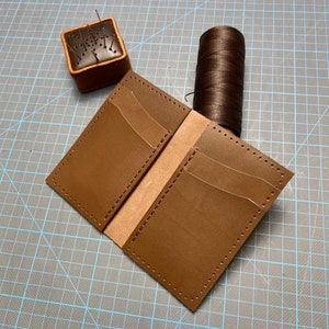 May include: Brown leather wallet with multiple card slots, being stitched together with brown thread. A spool of brown thread and a leather tool with needles are also visible.