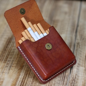 Könnte beinhalten: Eine braune Zigarettenetui aus Leder, geöffnet, um mehrere Zigaretten zu zeigen. Die Hülle hat einen Magnetverschluss und weiße Nähte an den Rändern. Das Leder hat ein reichhaltiges, strukturiertes Aussehen.