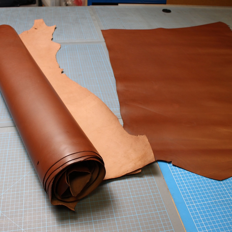 May include: A brown leather hide is rolled up and placed on a blue and gray cutting mat. A second piece of brown leather is laid flat on the mat.
