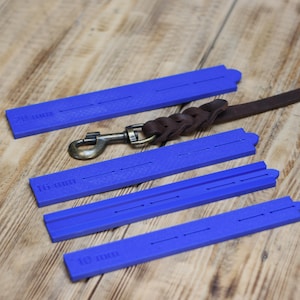 Puede incluir: Guías azules de plástico para correa de perro con medidas de 10 mm, 16 mm y 20 mm. También se muestra una correa de cuero marrón para perro con un cierre metálico. Los artículos están dispuestos sobre una superficie de madera.