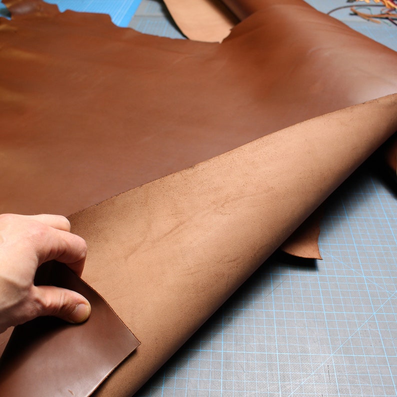 May include: A large piece of brown leather is folded in half, showing the inside of the leather. The leather is smooth and has a slight sheen. The leather is lying on a blue and white gridded surface.