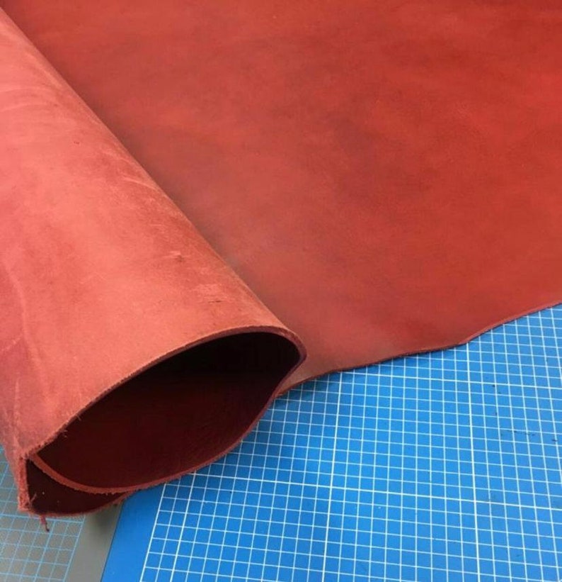 May include: A close-up of a piece of red leather rolled up on a blue cutting mat. The leather is smooth and has a slight sheen.