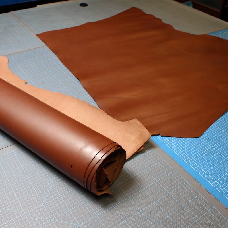 May include: A rolled piece of brown leather on a blue and gray cutting mat. A larger piece of brown leather is laid out on the mat.