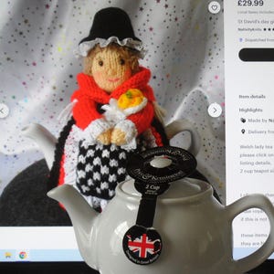 May include: A knitted Welsh lady tea cosy in red, white and black, with a white lace collar and a white flower. The cosy is on a grey teapot with a 2-cup label. The teapot is on a white surface.