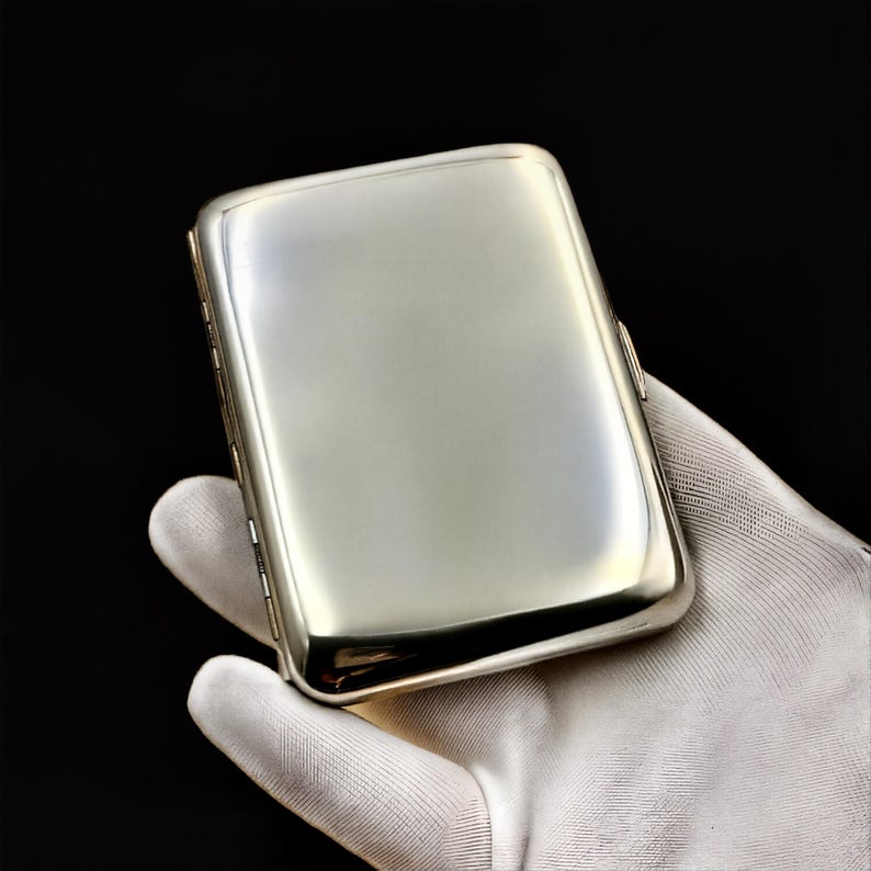 May include: A silver cigarette case with a smooth, polished finish. The case is closed and has a rectangular shape.