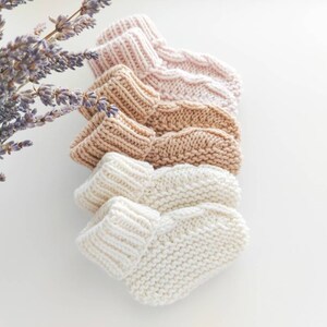 May include: A stack of four knitted baby booties in varying neutral colors: pale pink, tan, light brown, and cream. The booties are arranged in a row, showcasing their textured knit design. A sprig of lavender is in the upper left corner.