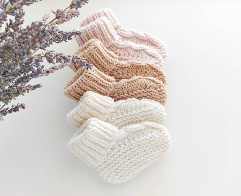 May include: A stack of four knitted baby booties in varying neutral colors: pale pink, tan, light brown, and cream. The booties are arranged in a row, showcasing their textured knit design. A sprig of lavender is in the upper left corner.